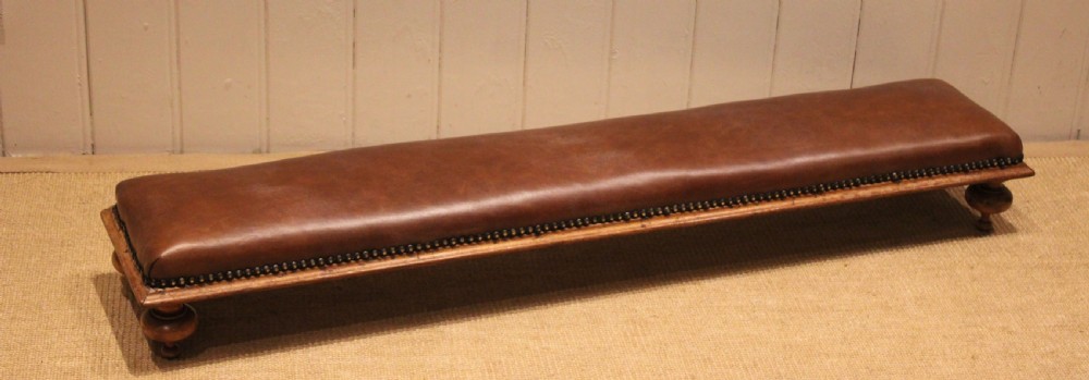 A long, low Victorian footstool upholstered in warm brown leather with brass studding along the lower edge, supported by short turned walnut feet. View VI
