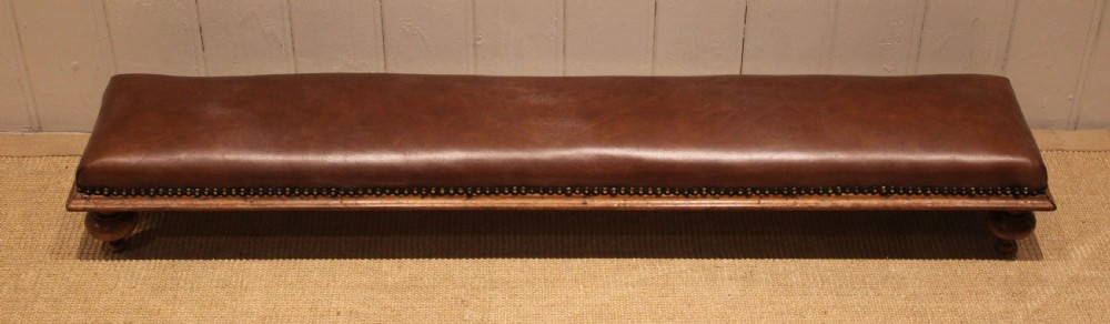 A long, low Victorian footstool upholstered in warm brown leather with brass studding along the lower edge, supported by short turned walnut feet. View VII