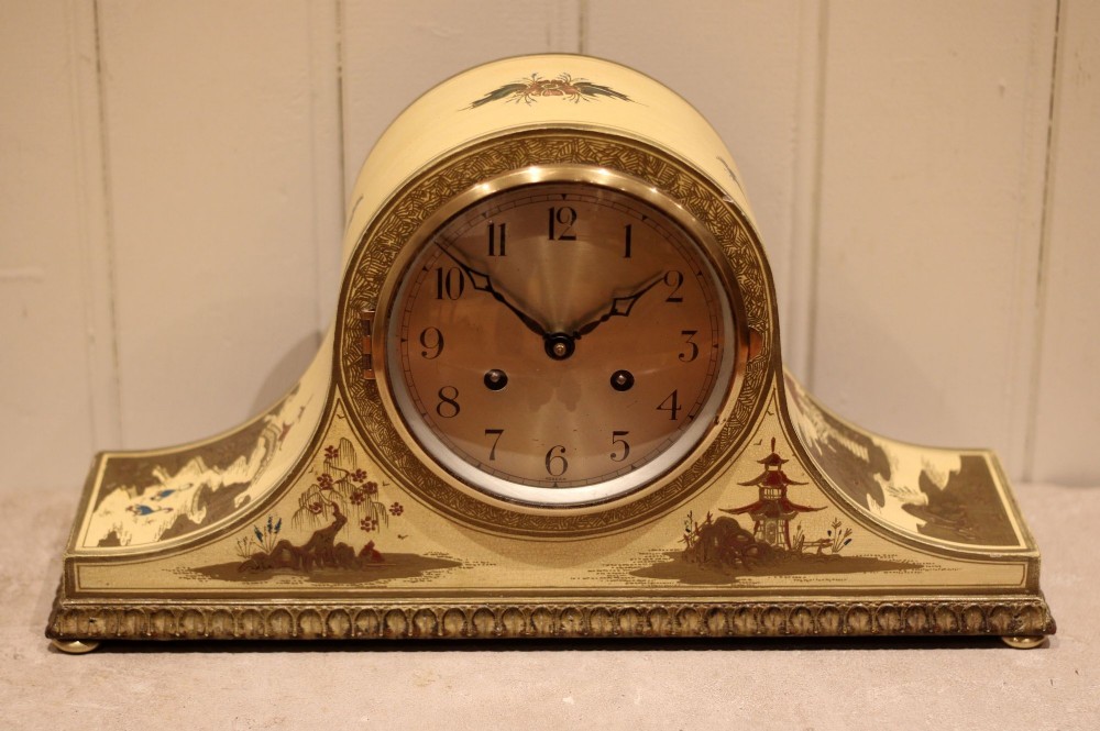 An arched cream lacquered mantel clock featuring hand-painted Chinoiserie pagoda landscapes and a brass-rimmed dial with Arabic numerals.