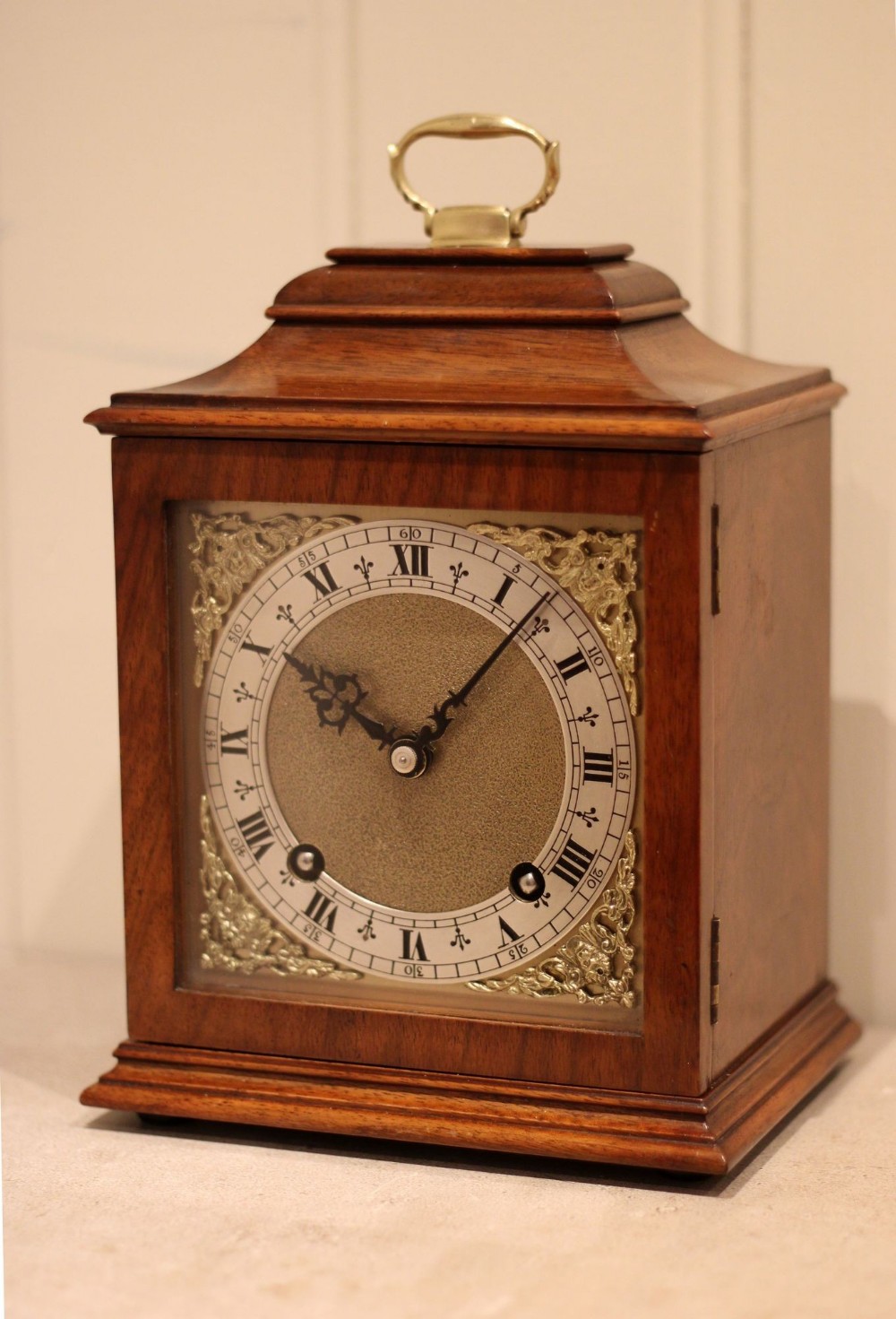 A walnut bracket clock featuring a brass carrying handle on top, a clear glass door, and a silvered dial with black Roman numerals and ornate brass spandrels.