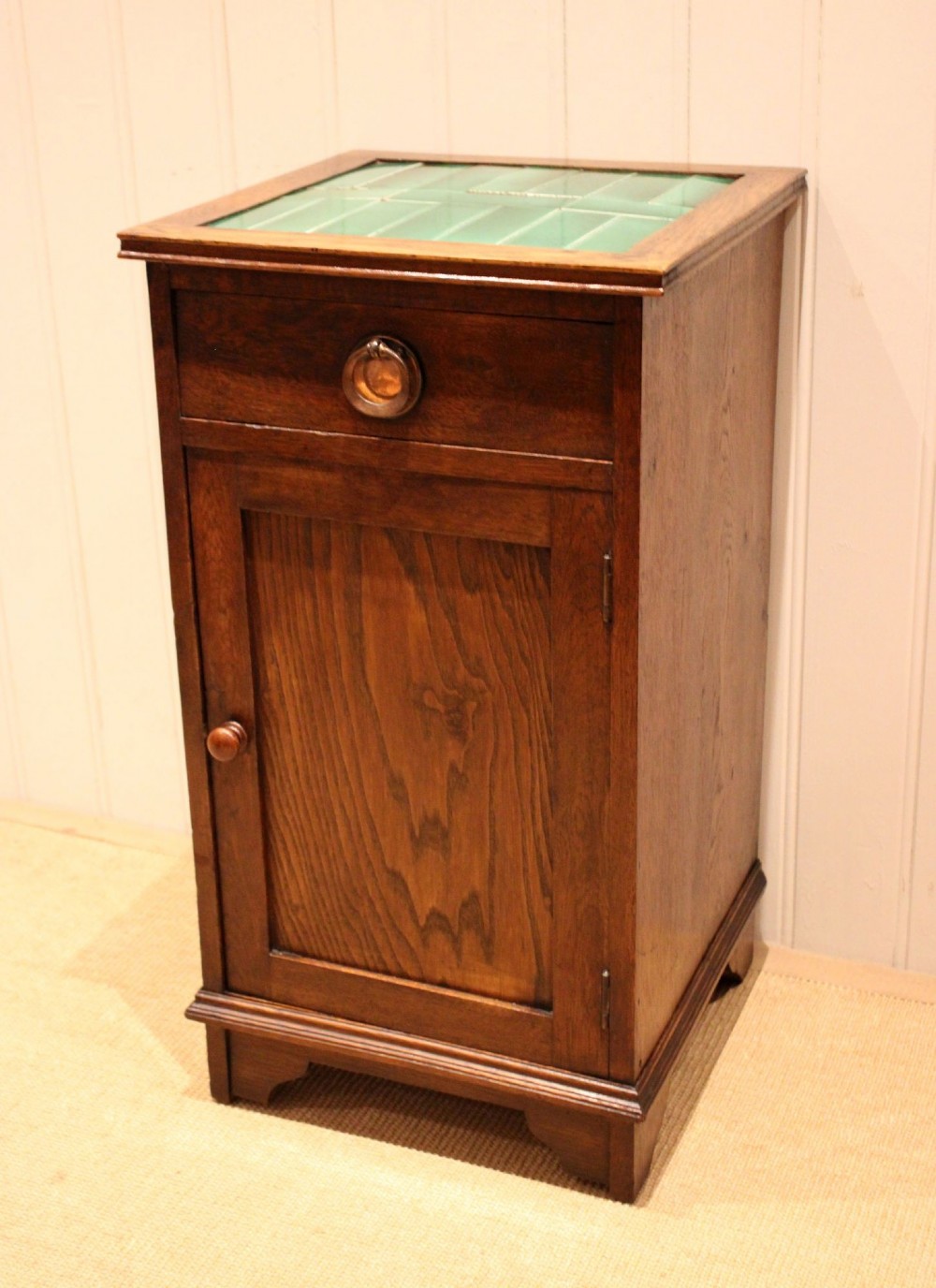 An Arts and Crafts oak cabinet featuring a recessed green glazed tile top, a single drawer with a round ring pull, and a lower panelled cupboard door.