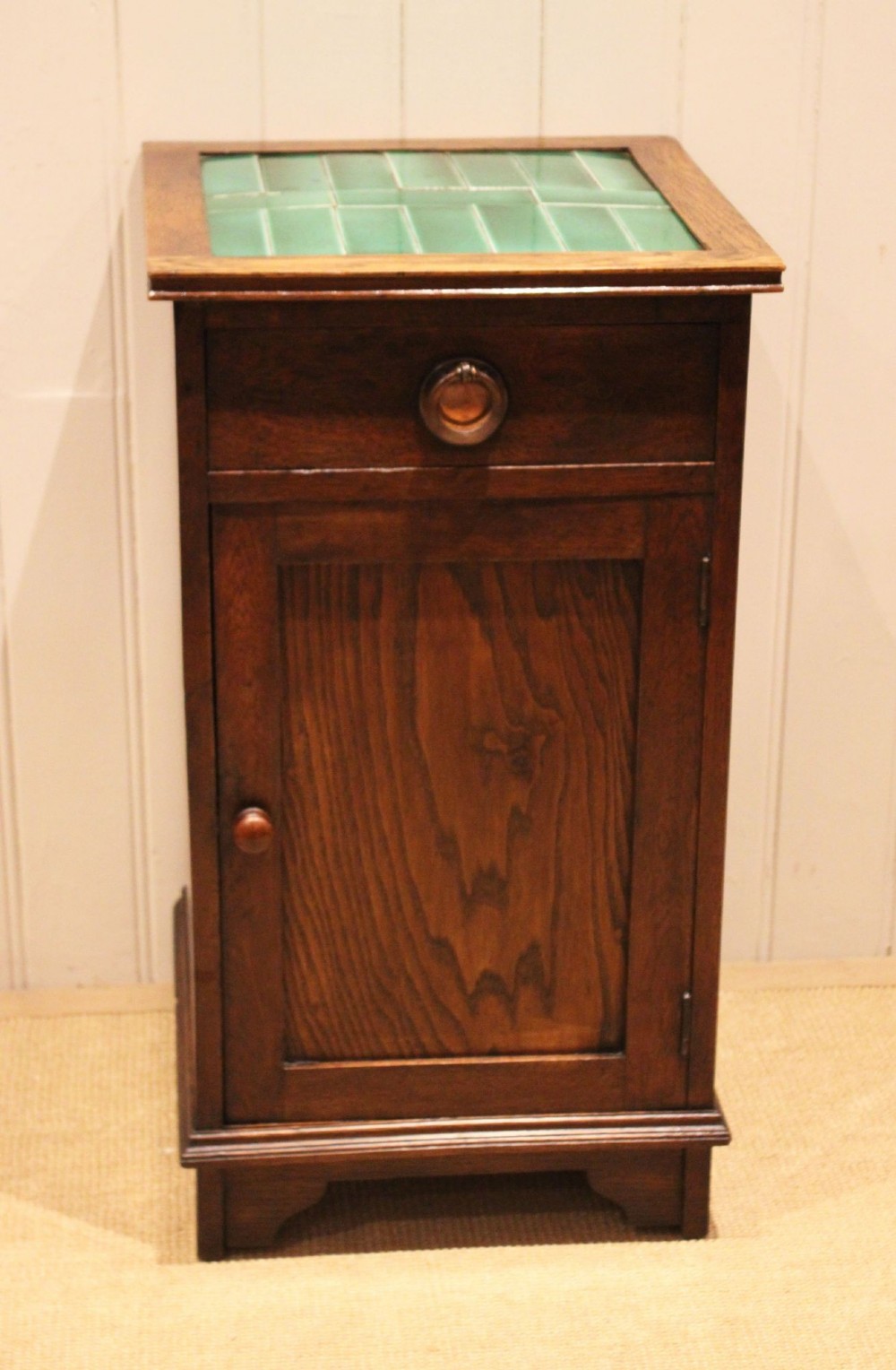 An Arts and Crafts oak cabinet featuring a recessed green glazed tile top, a single drawer with a round ring pull, and a lower panelled cupboard door. View II