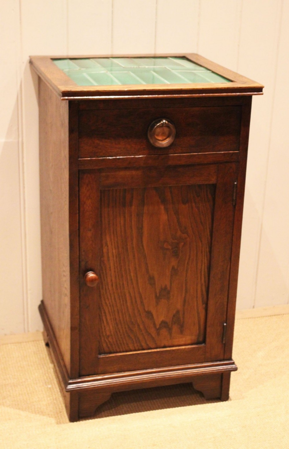 An Arts and Crafts oak cabinet featuring a recessed green glazed tile top, a single drawer with a round ring pull, and a lower panelled cupboard door. View III
