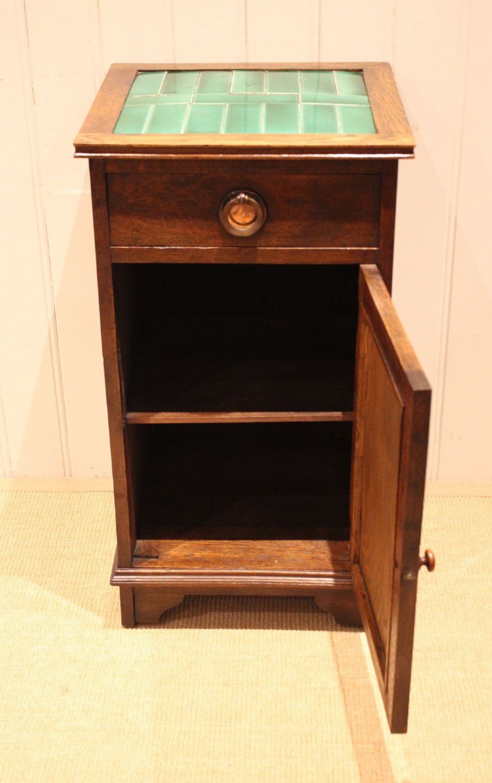 An Arts and Crafts oak cabinet featuring a recessed green glazed tile top, a single drawer with a round ring pull, and a lower panelled cupboard door. View IV