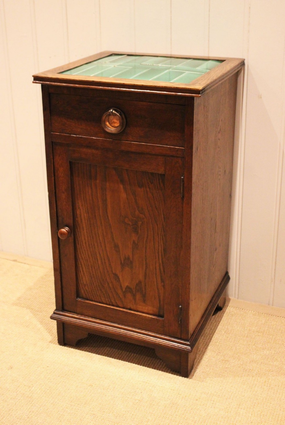 An Arts and Crafts oak cabinet featuring a recessed green glazed tile top, a single drawer with a round ring pull, and a lower panelled cupboard door. View VI