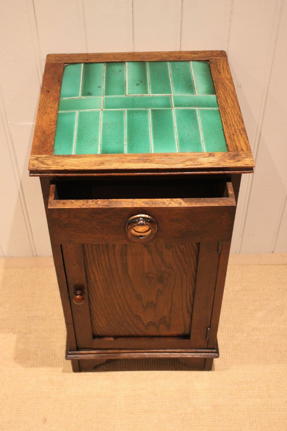 An Arts and Crafts oak cabinet featuring a recessed green glazed tile top, a single drawer with a round ring pull, and a lower panelled cupboard door. View VII