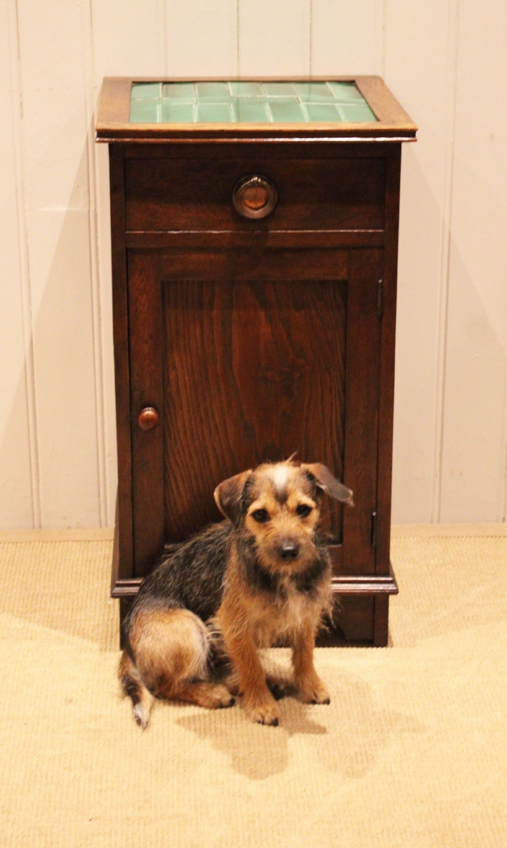 An Arts and Crafts oak cabinet featuring a recessed green glazed tile top, a single drawer with a round ring pull, and a lower panelled cupboard door. View VIII