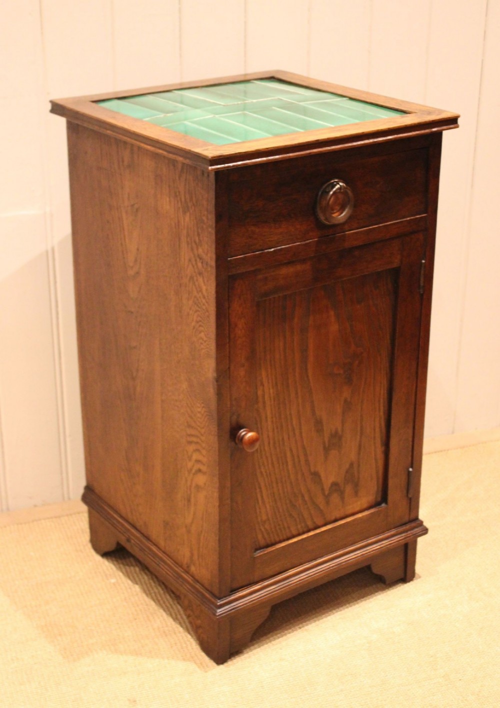 An Arts and Crafts oak cabinet featuring a recessed green glazed tile top, a single drawer with a round ring pull, and a lower panelled cupboard door. View X