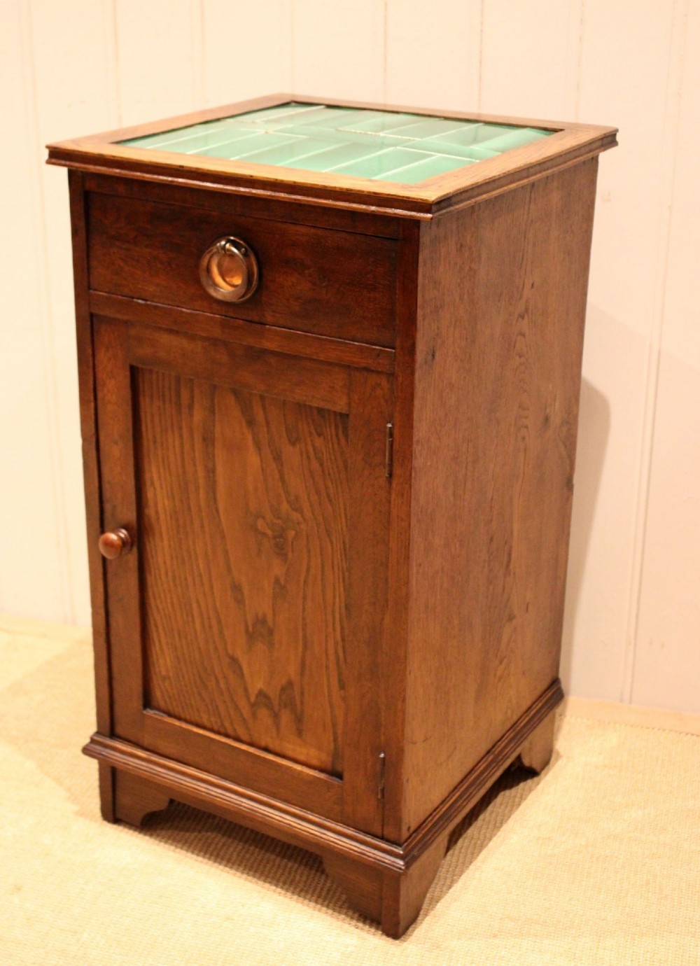 An Arts and Crafts oak cabinet featuring a recessed green glazed tile top, a single drawer with a round ring pull, and a lower panelled cupboard door. View XI
