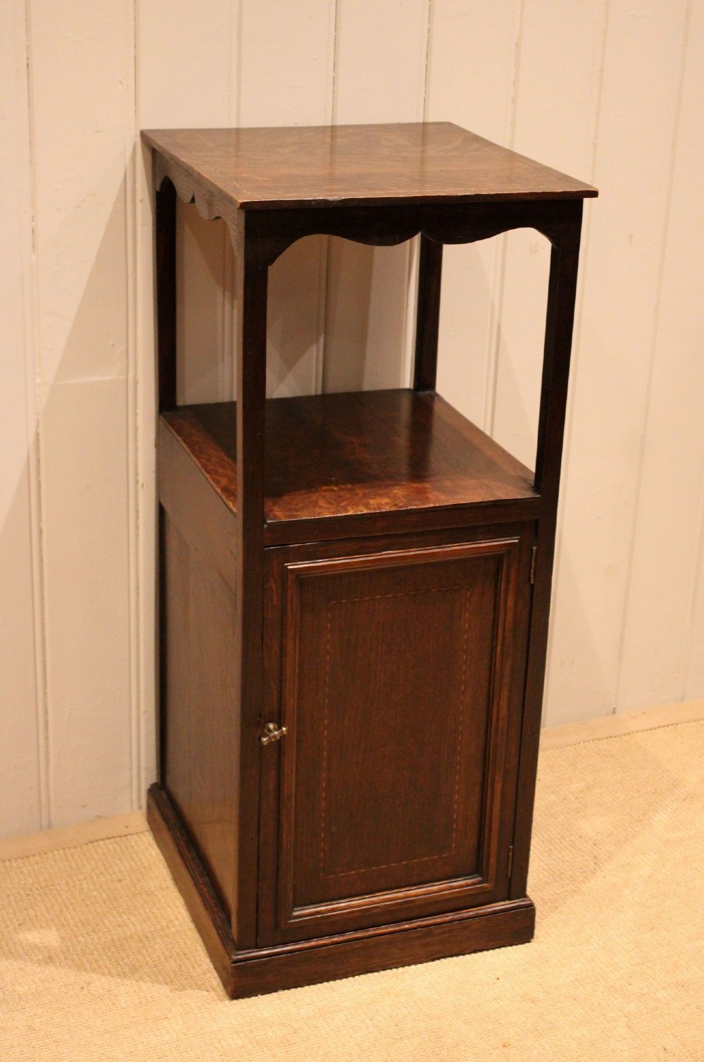 A dark oak Edwardian bedside cabinet with an open upper shelf, shaped apron, and a panelled single door below. View IX