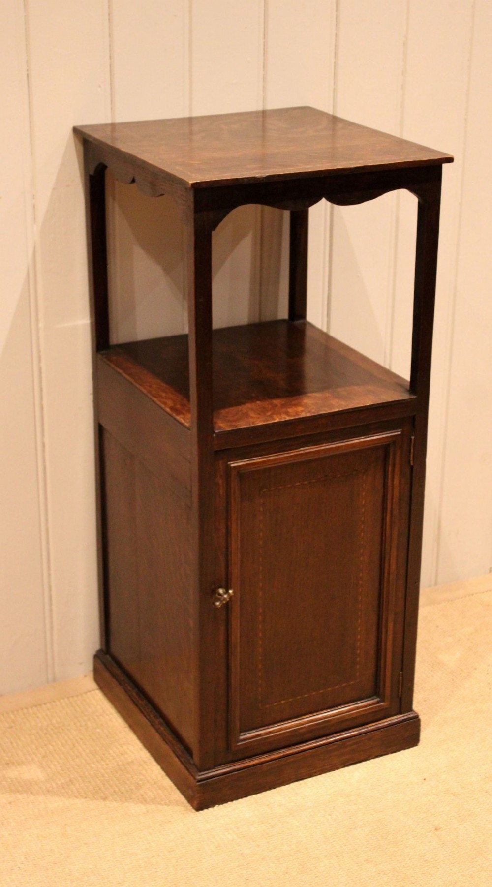 A dark oak Edwardian bedside cabinet with an open upper shelf, shaped apron, and a panelled single door below. View X