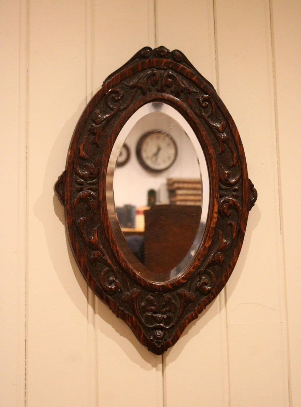 A small oval wall mirror featuring a heavily carved dark oak wooden frame with scroll motifs and a bevelled glass plate.