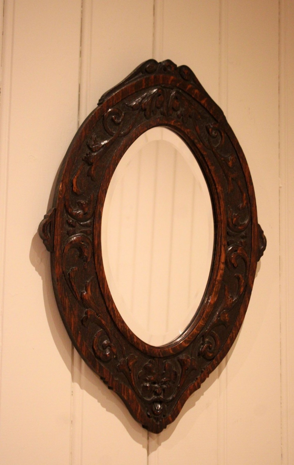 A small oval wall mirror featuring a heavily carved dark oak wooden frame with scroll motifs and a bevelled glass plate. View II