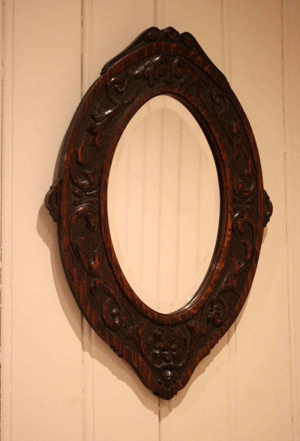 A small oval wall mirror featuring a heavily carved dark oak wooden frame with scroll motifs and a bevelled glass plate. View IV