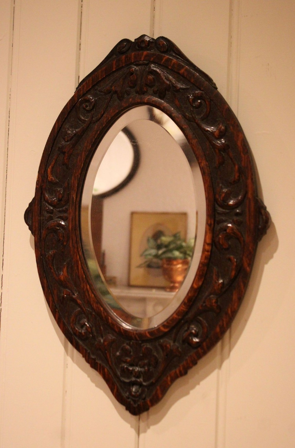 A small oval wall mirror featuring a heavily carved dark oak wooden frame with scroll motifs and a bevelled glass plate. View V