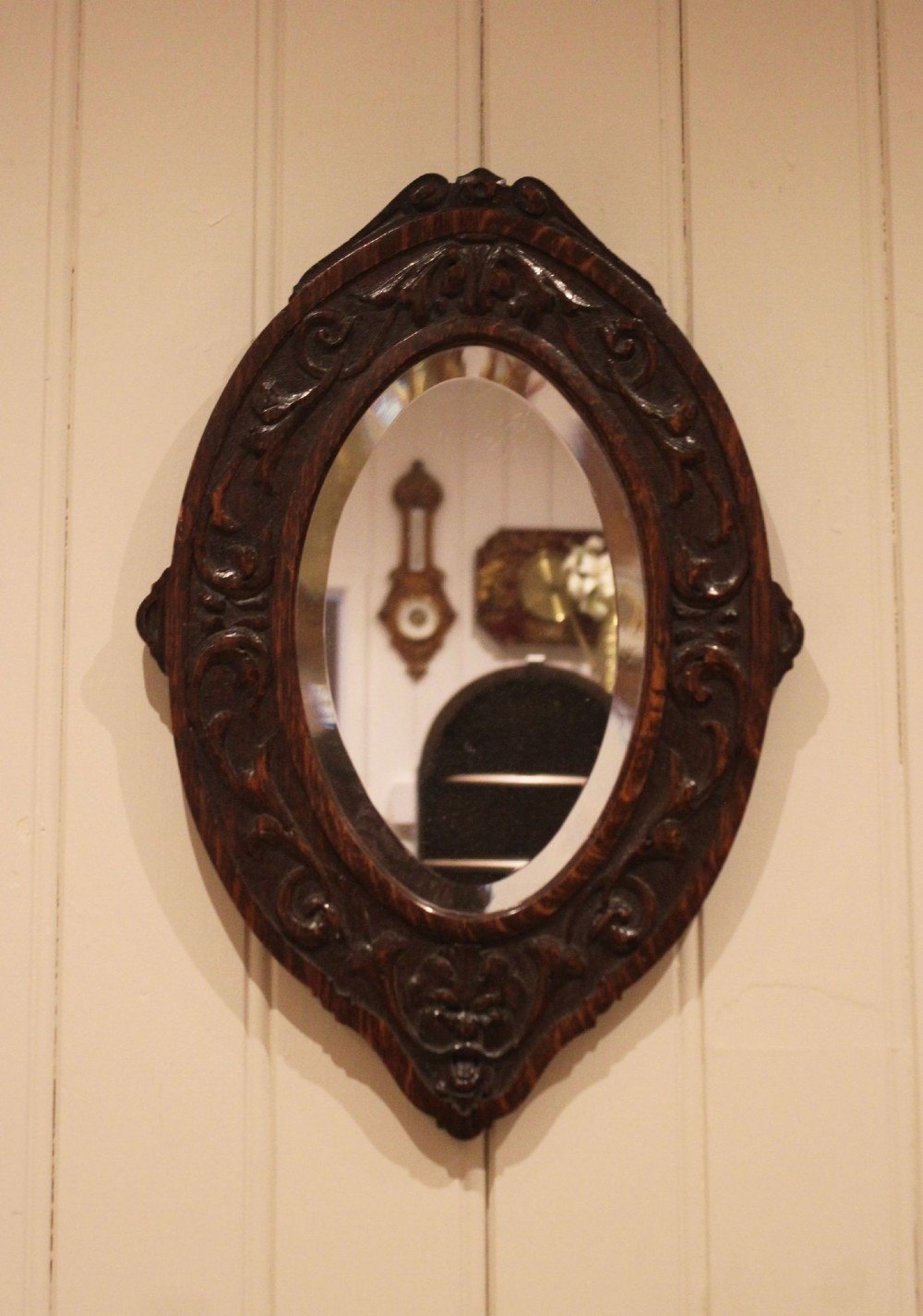 A small oval wall mirror featuring a heavily carved dark oak wooden frame with scroll motifs and a bevelled glass plate. View VII