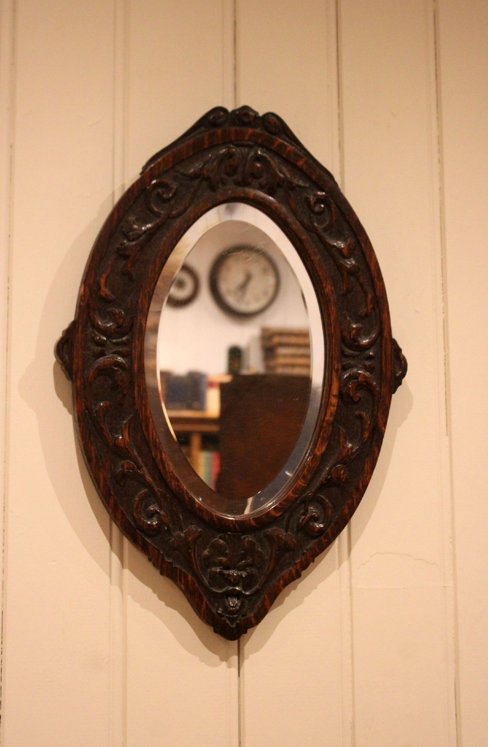 A small oval wall mirror featuring a heavily carved dark oak wooden frame with scroll motifs and a bevelled glass plate. View IX