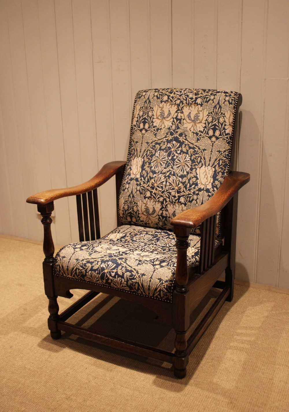 An Arts and Crafts oak reclining armchair featuring slatted sides, turned front legs, wide armrests, and floral pattern upholstery in blue and cream tones. View X