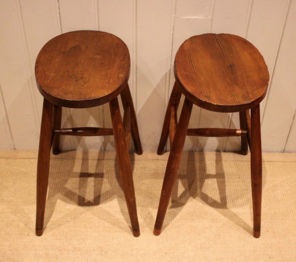 A pair of rustic warm brown wooden stools crafted with oval seats, turned splayed legs, and supporting stretchers. View X