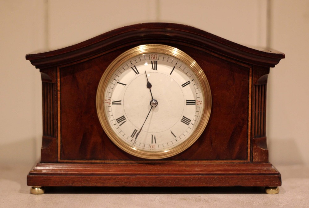 An Edwardian mahogany mantel clock featuring an arched wooden case, brass bezel, white circular dial with Roman numerals, and brass feet.