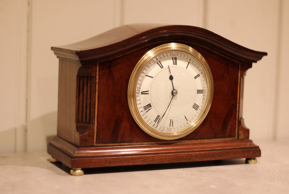 An Edwardian mahogany mantel clock featuring an arched wooden case, brass bezel, white circular dial with Roman numerals, and brass feet. View II