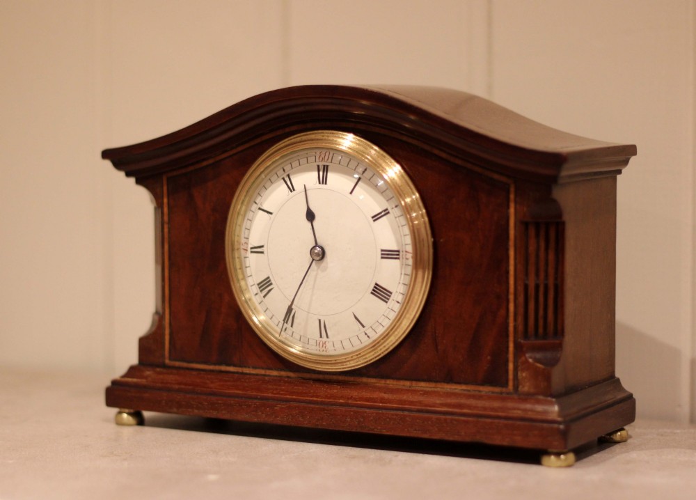 An Edwardian mahogany mantel clock featuring an arched wooden case, brass bezel, white circular dial with Roman numerals, and brass feet. View III
