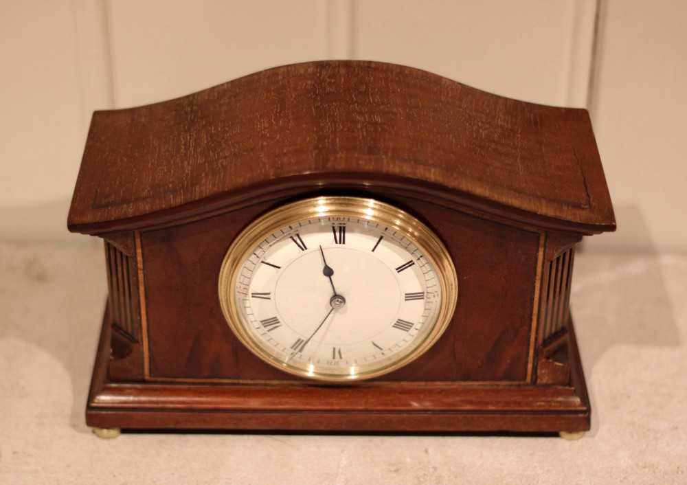 An Edwardian mahogany mantel clock featuring an arched wooden case, brass bezel, white circular dial with Roman numerals, and brass feet. View IV