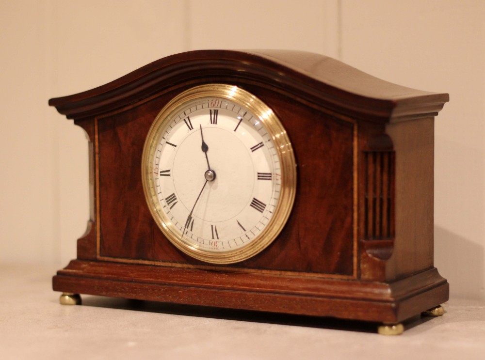 An Edwardian mahogany mantel clock featuring an arched wooden case, brass bezel, white circular dial with Roman numerals, and brass feet. View V