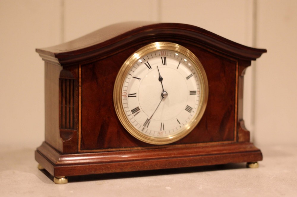 An Edwardian mahogany mantel clock featuring an arched wooden case, brass bezel, white circular dial with Roman numerals, and brass feet. View VI