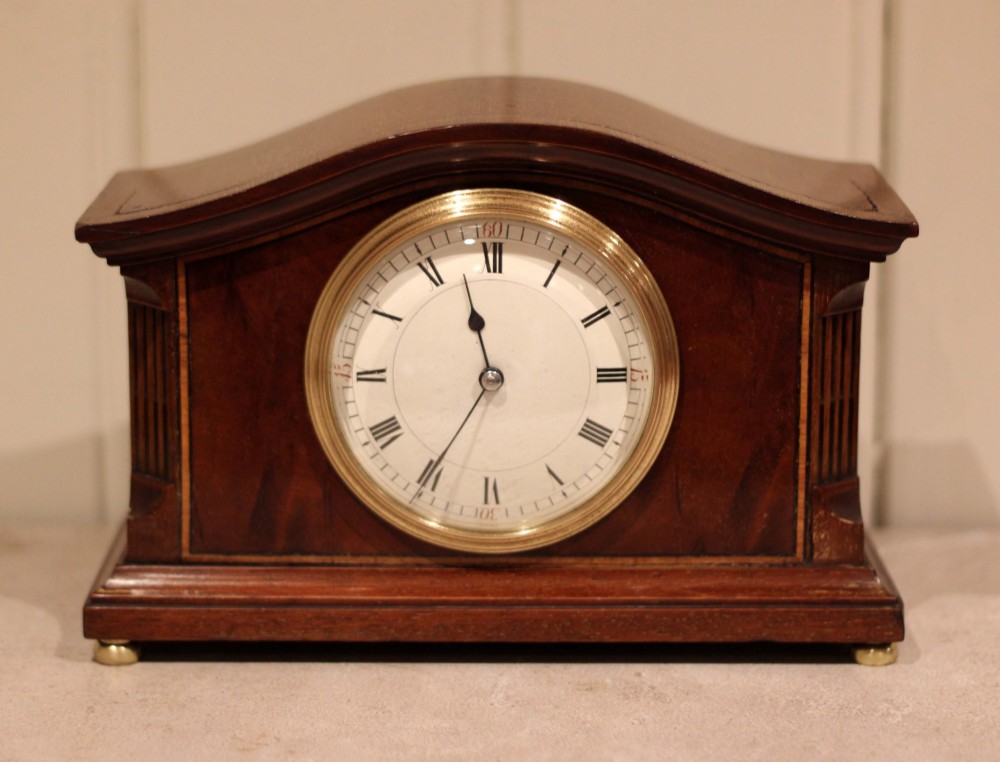 An Edwardian mahogany mantel clock featuring an arched wooden case, brass bezel, white circular dial with Roman numerals, and brass feet. View VII