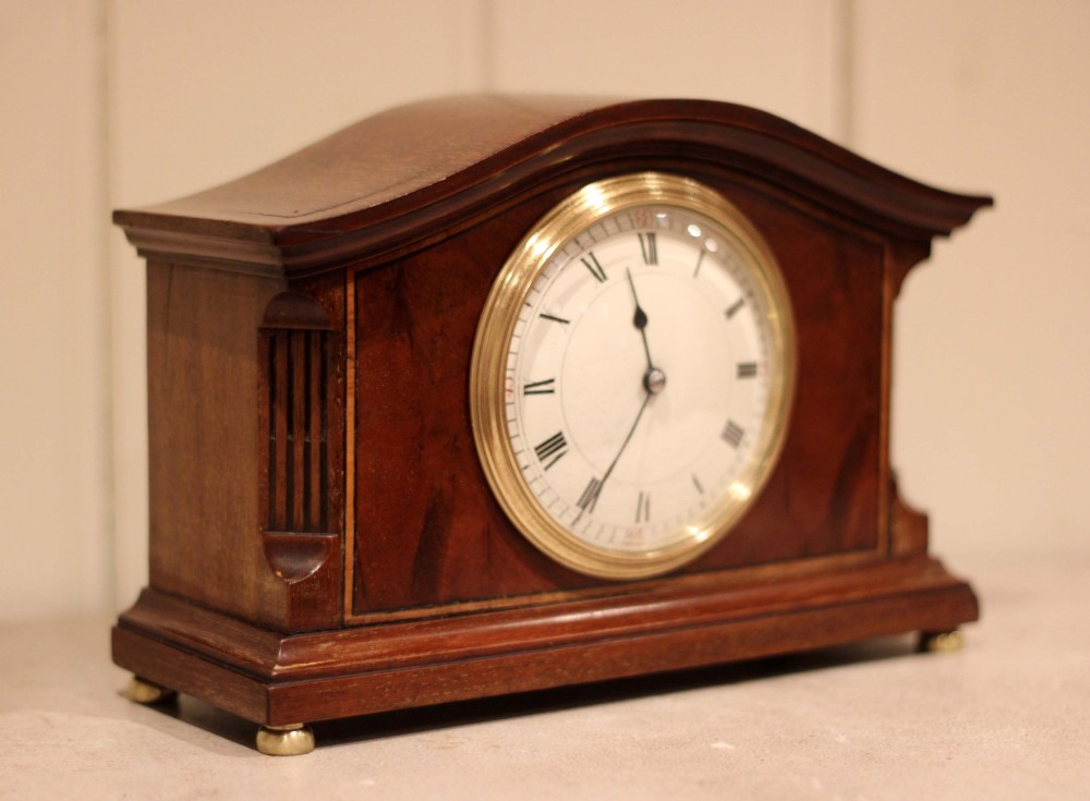 An Edwardian mahogany mantel clock featuring an arched wooden case, brass bezel, white circular dial with Roman numerals, and brass feet. View VIII
