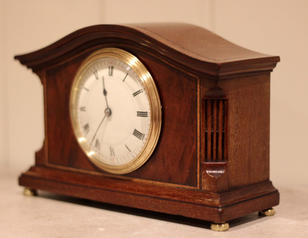 An Edwardian mahogany mantel clock featuring an arched wooden case, brass bezel, white circular dial with Roman numerals, and brass feet. View IX