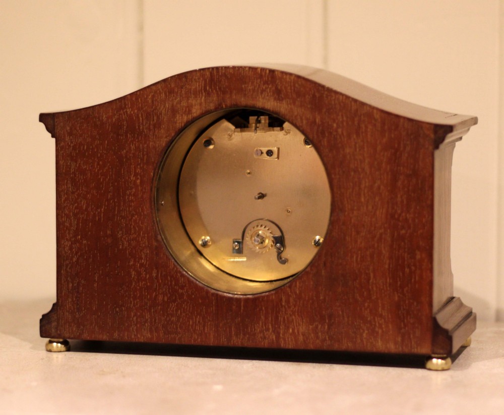 An Edwardian mahogany mantel clock featuring an arched wooden case, brass bezel, white circular dial with Roman numerals, and brass feet. View X