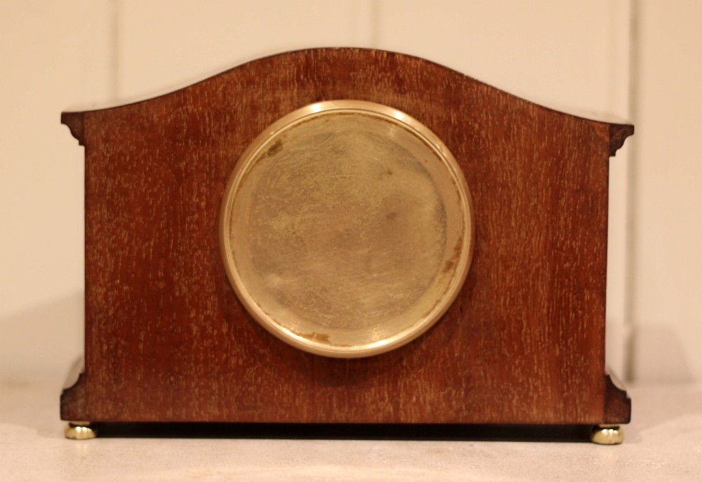 An Edwardian mahogany mantel clock featuring an arched wooden case, brass bezel, white circular dial with Roman numerals, and brass feet. View XII