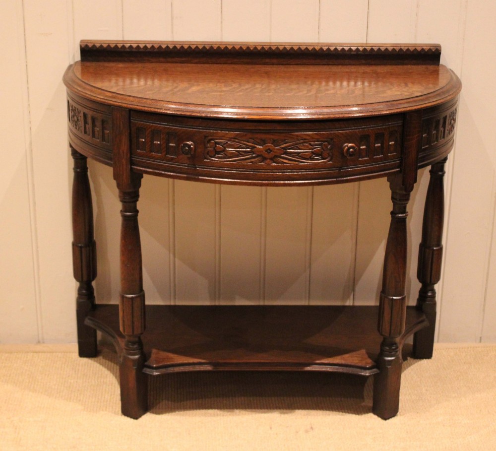 A dark carved oak demilune console table featuring a semi-circular top, carved apron, turned front legs, and a lower shelf.