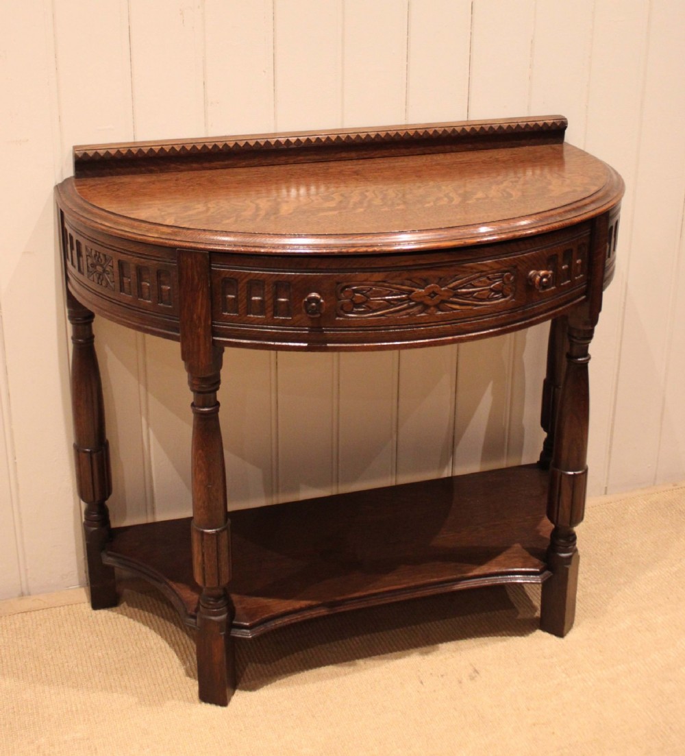 A dark carved oak demilune console table featuring a semi-circular top, carved apron, turned front legs, and a lower shelf. View II