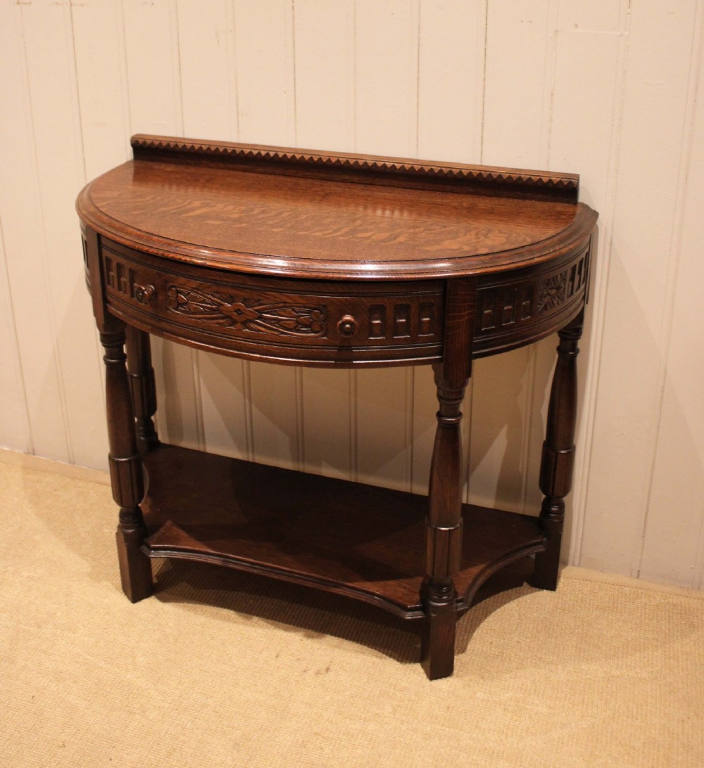A dark carved oak demilune console table featuring a semi-circular top, carved apron, turned front legs, and a lower shelf. View III