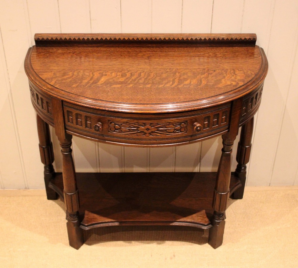 A dark carved oak demilune console table featuring a semi-circular top, carved apron, turned front legs, and a lower shelf. View IV