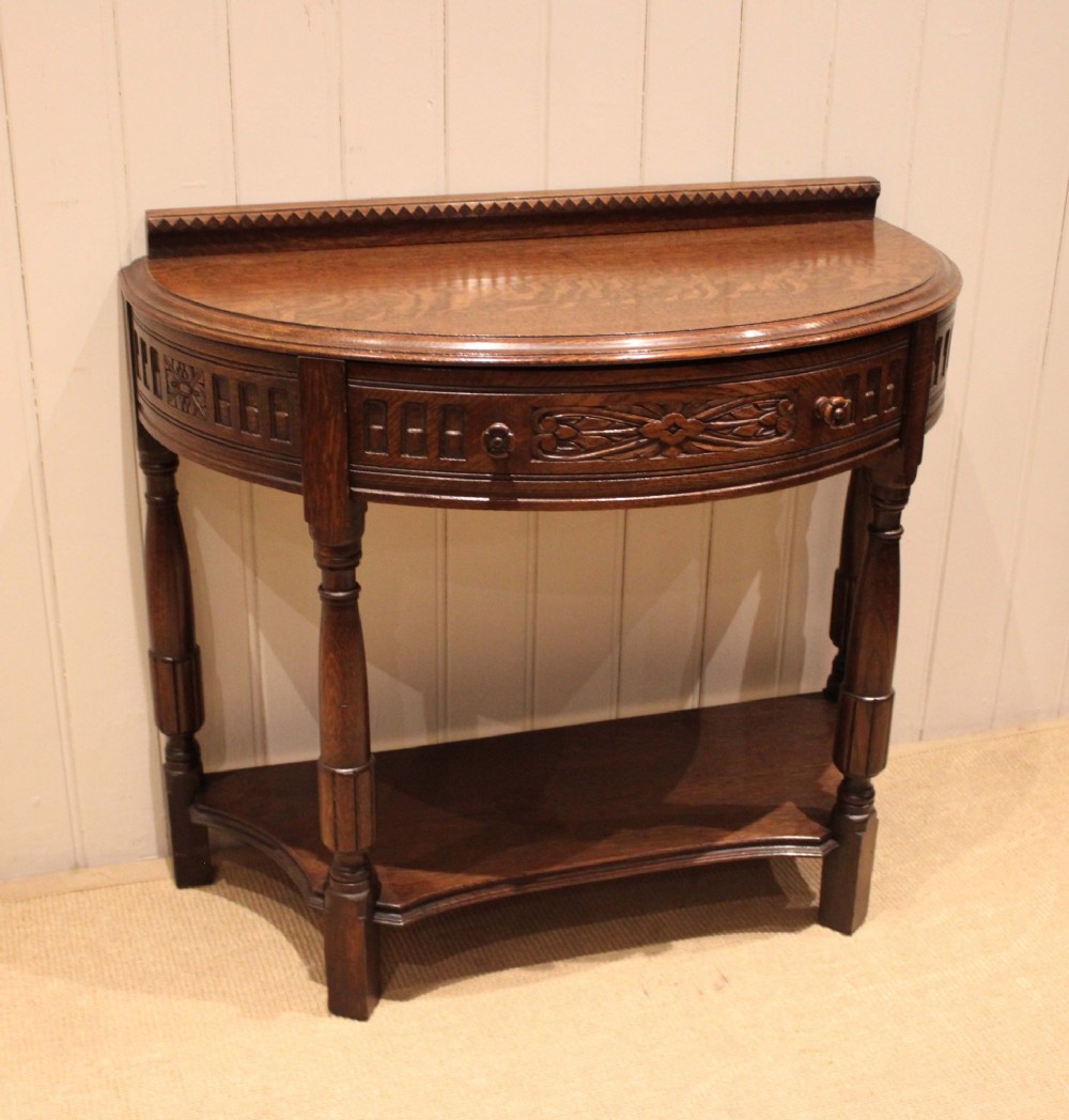 A dark carved oak demilune console table featuring a semi-circular top, carved apron, turned front legs, and a lower shelf. View V