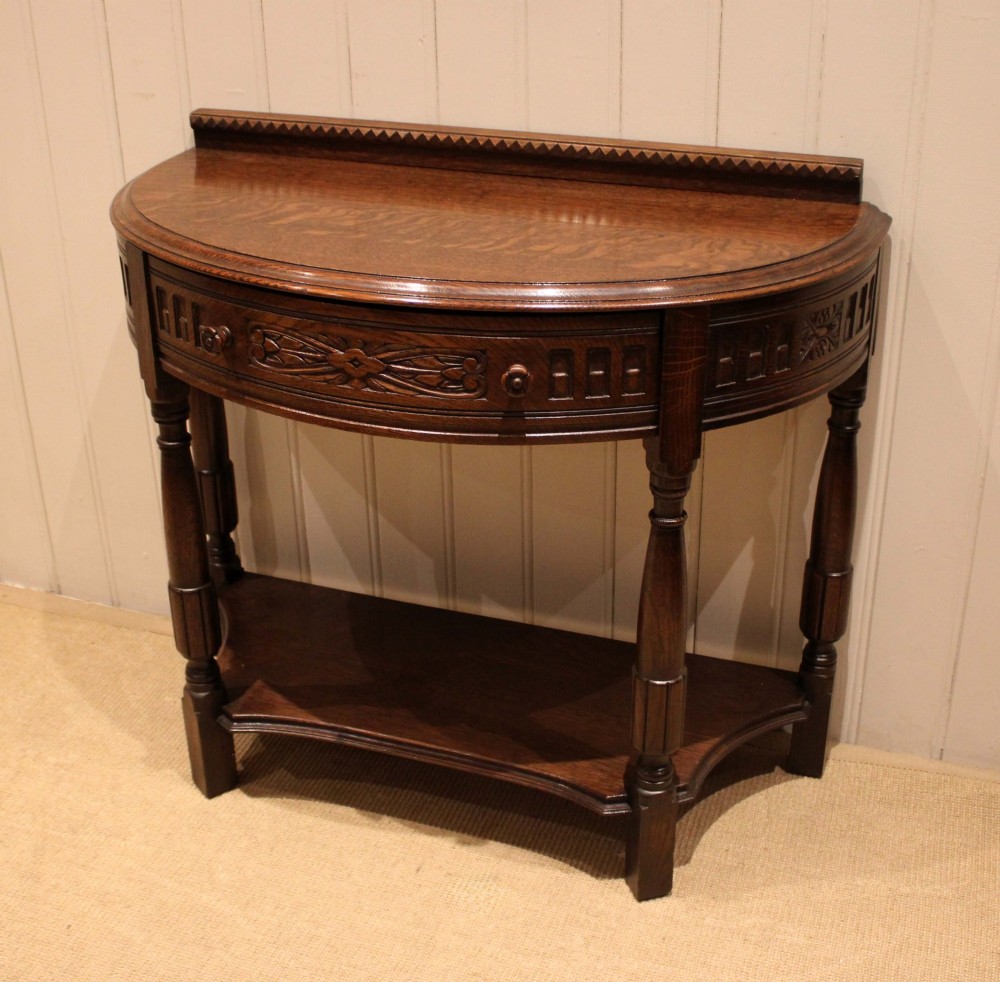 A dark carved oak demilune console table featuring a semi-circular top, carved apron, turned front legs, and a lower shelf. View VI
