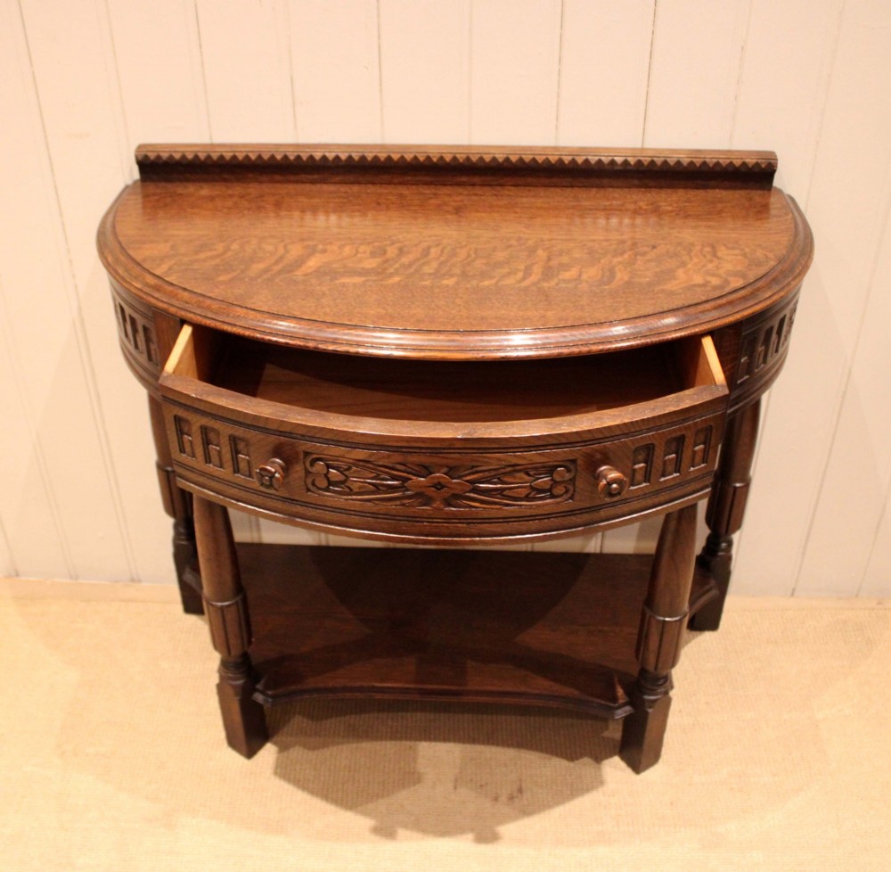 A dark carved oak demilune console table featuring a semi-circular top, carved apron, turned front legs, and a lower shelf. View VII
