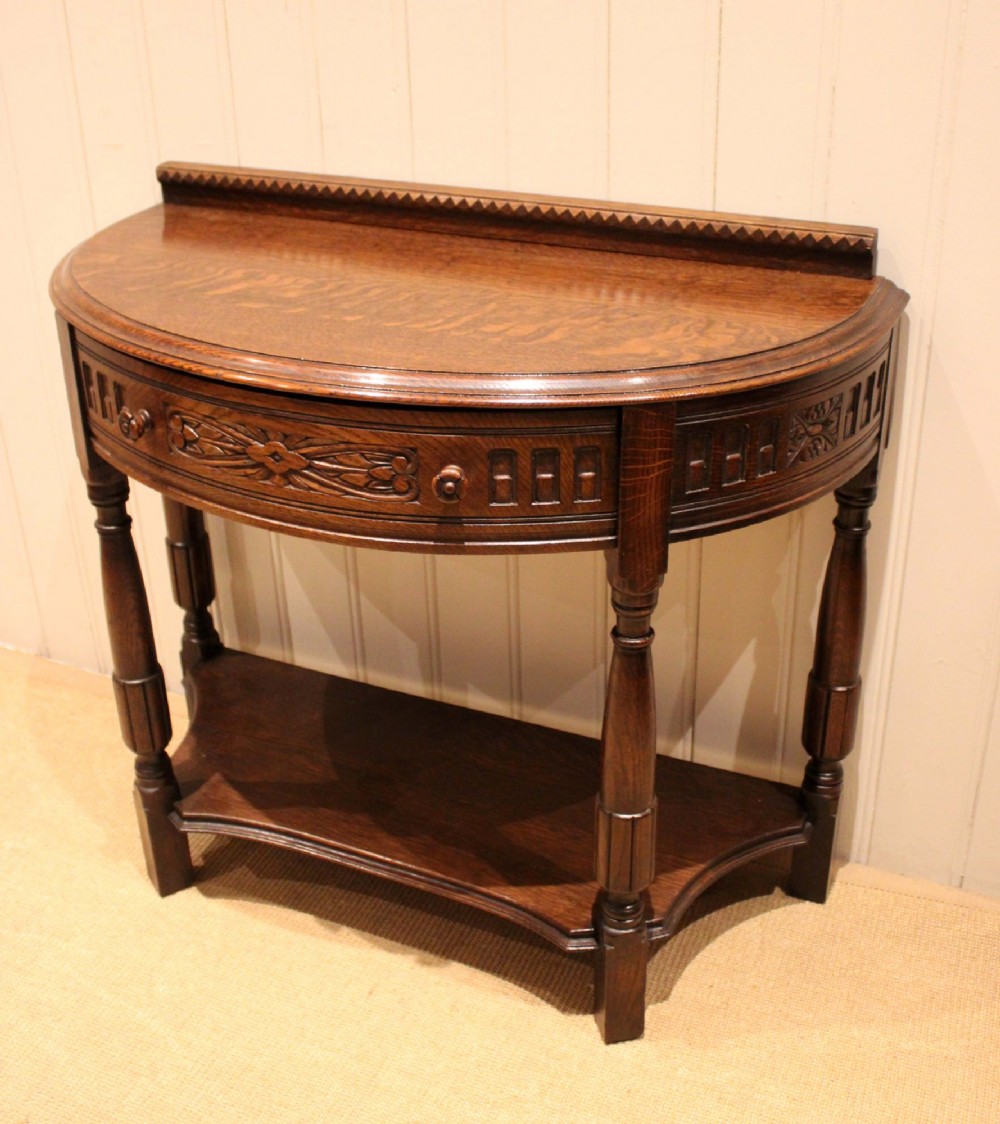 A dark carved oak demilune console table featuring a semi-circular top, carved apron, turned front legs, and a lower shelf. View VIII