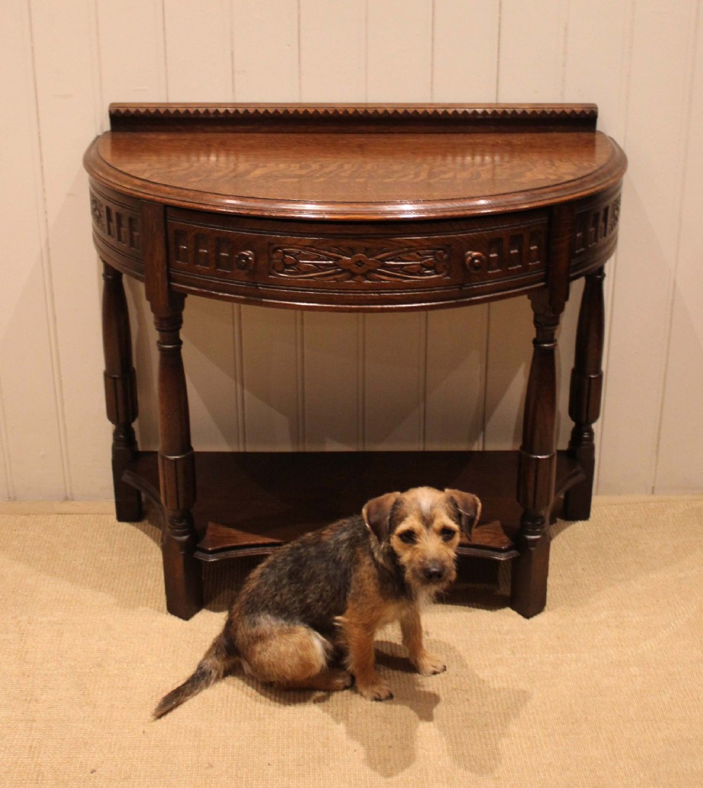 A dark carved oak demilune console table featuring a semi-circular top, carved apron, turned front legs, and a lower shelf. View IX