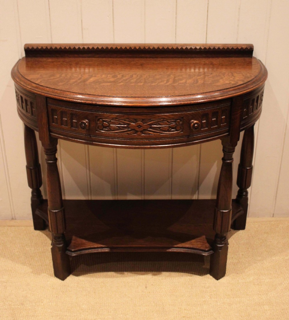 A dark carved oak demilune console table featuring a semi-circular top, carved apron, turned front legs, and a lower shelf. View X