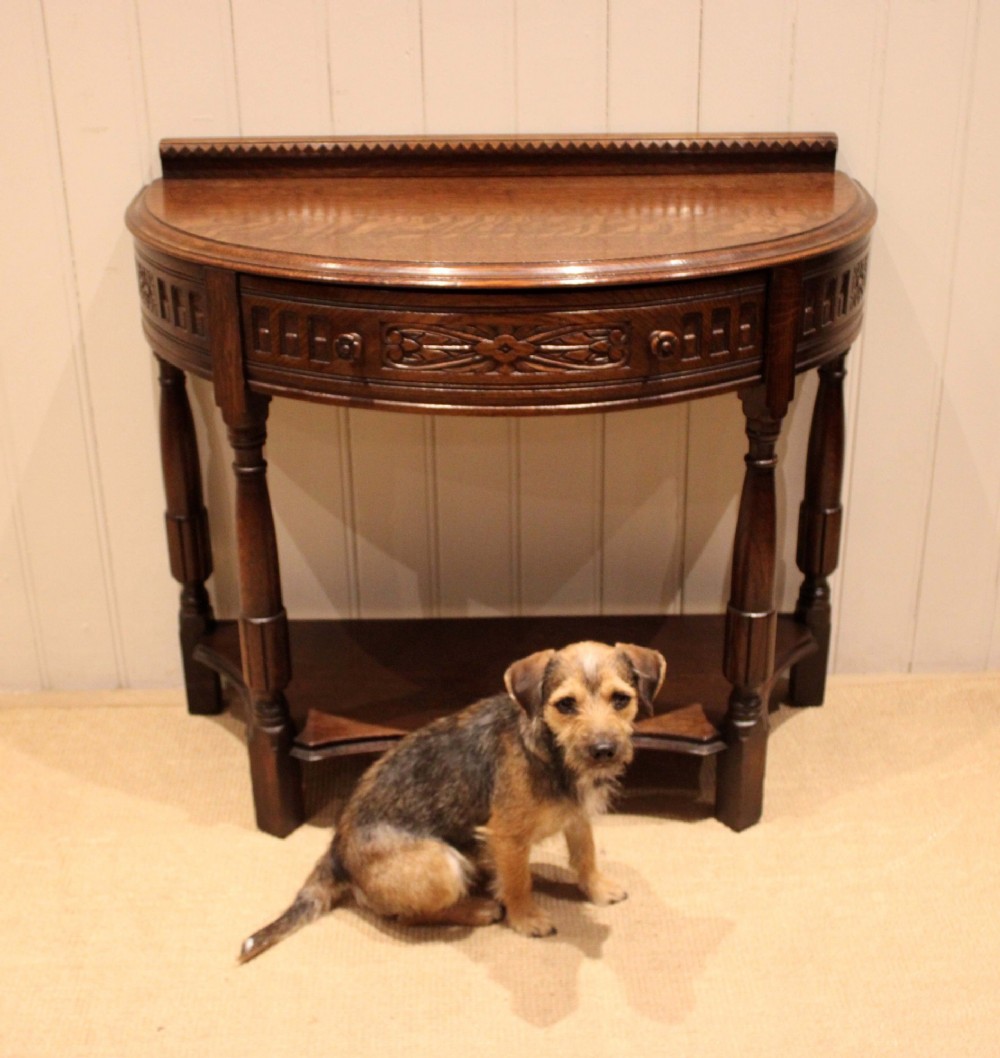 A dark carved oak demilune console table featuring a semi-circular top, carved apron, turned front legs, and a lower shelf. View XI
