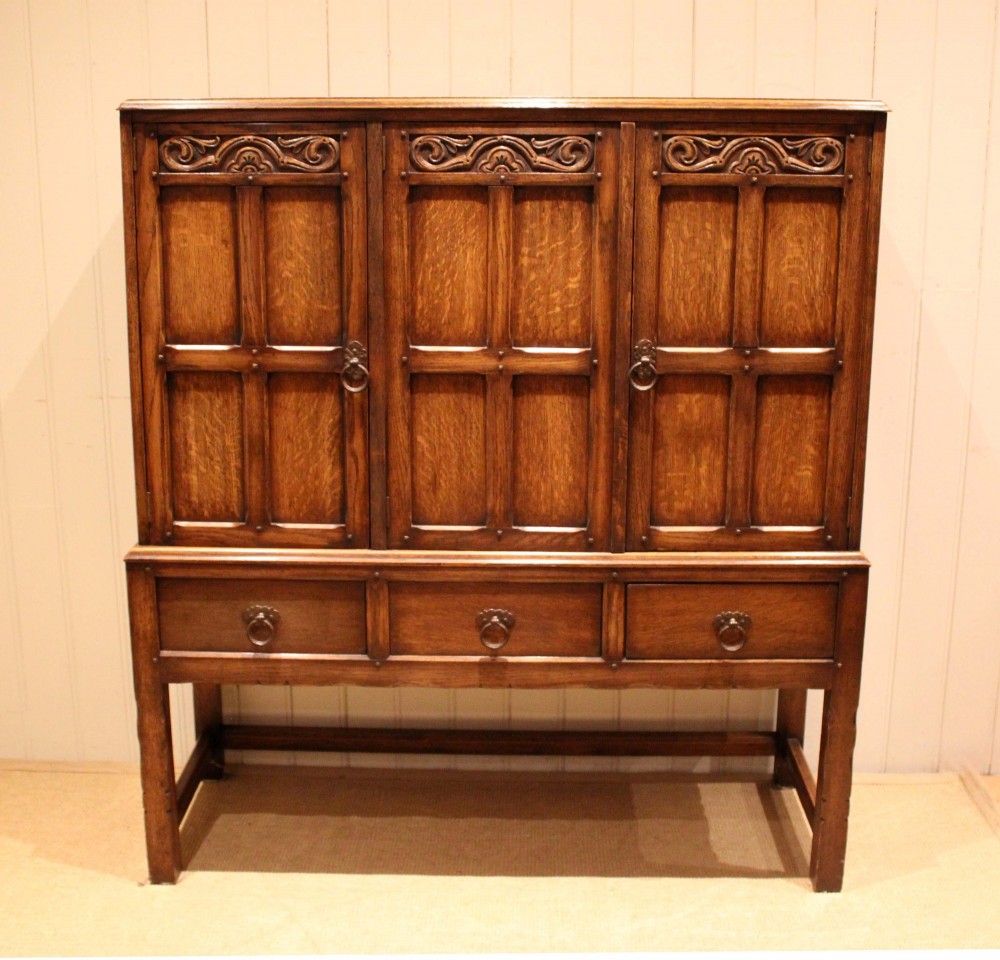 A solid oak side cabinet featuring three carved panel doors above three short drawers on square legs.