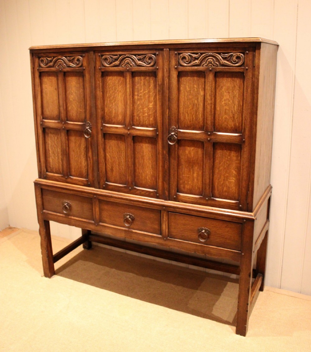A solid oak side cabinet featuring three carved panel doors above three short drawers on square legs. View II