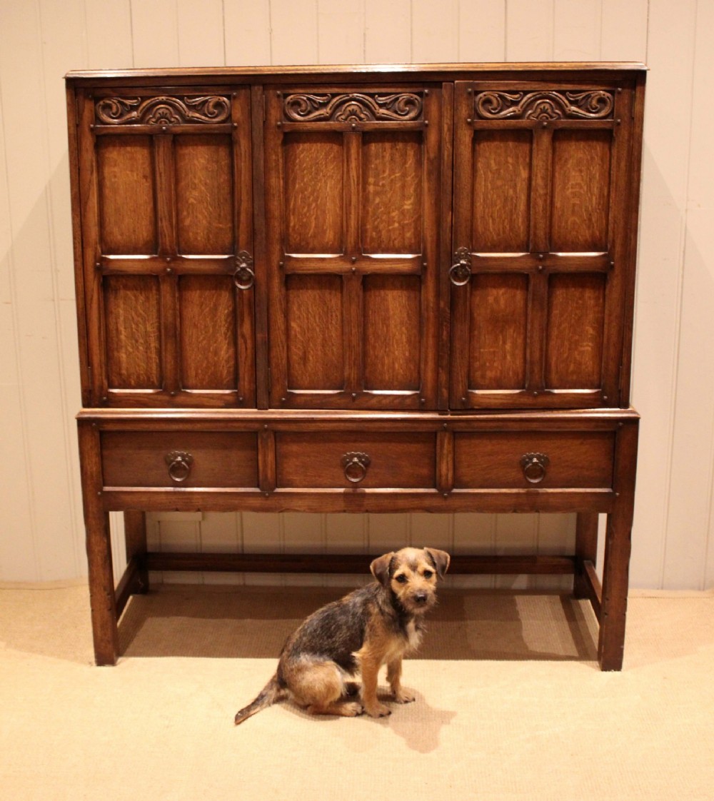 A solid oak side cabinet featuring three carved panel doors above three short drawers on square legs. View X
