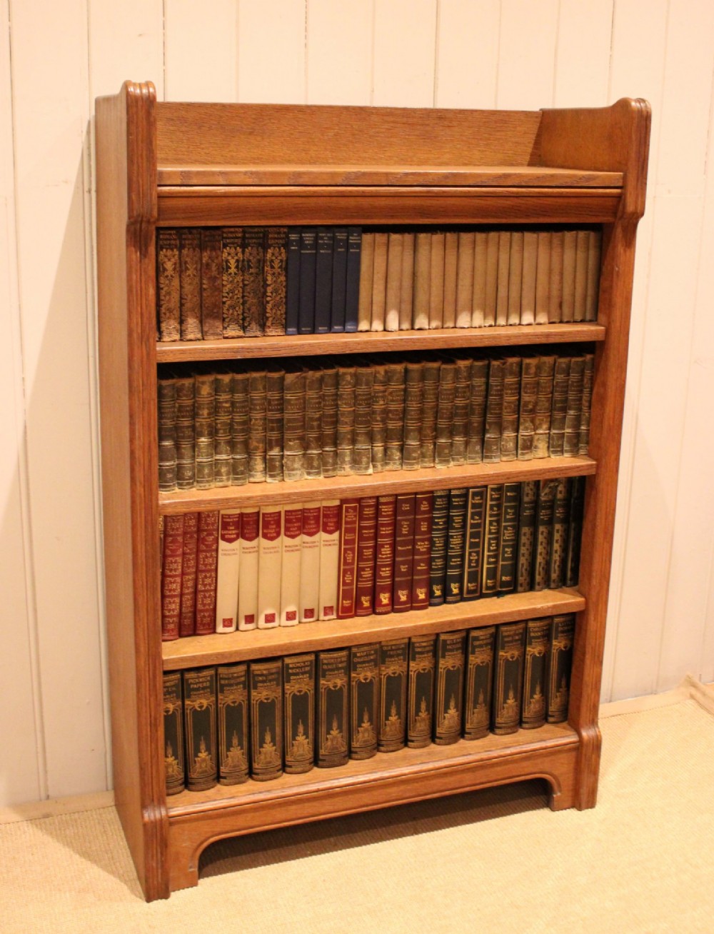 A freestanding light oak open bookcase featuring four horizontal shelves, a raised gallery rail, and curved side brackets.