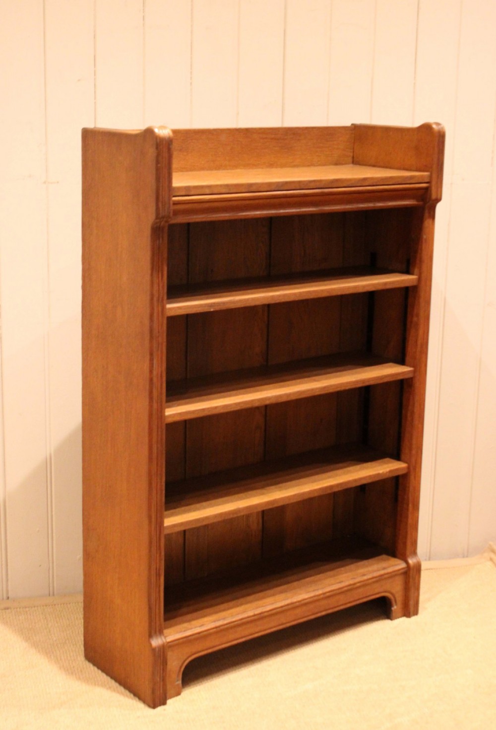 A freestanding light oak open bookcase featuring four horizontal shelves, a raised gallery rail, and curved side brackets. View X